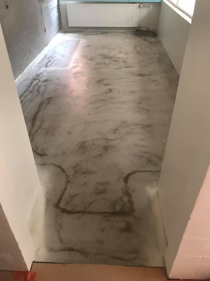 Huoneen betonilattia, jossa on epätasainen pinta ja valkoinen patteri seinällä.