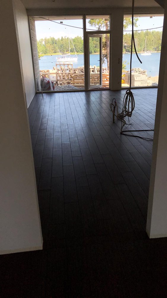 Sisätila, jossa on tumma puulattia, suuret ikkunat, joista näkyy järvimaisema veneineen ja puupinoja ulkona.