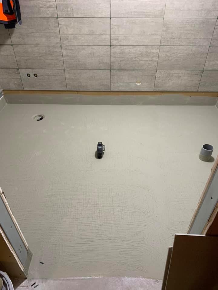 Valokuva kylpyhuoneesta, jossa on harmaat laatat seinillä ja tasainen, harmaa lattia, jossa on viemäri ja pieni vesivaaka.