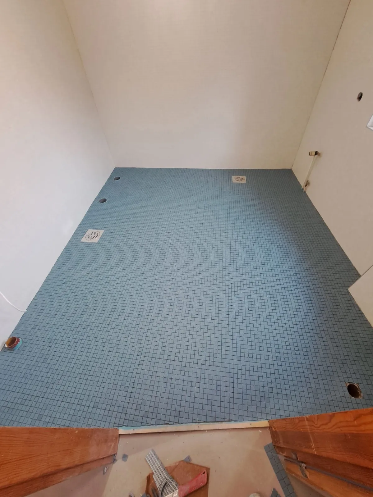 Korkea kuvakulma kylpyhuoneesta, jossa on siniset mosaiikkilaatat, valkoiset seinät ja puinen kynnys.