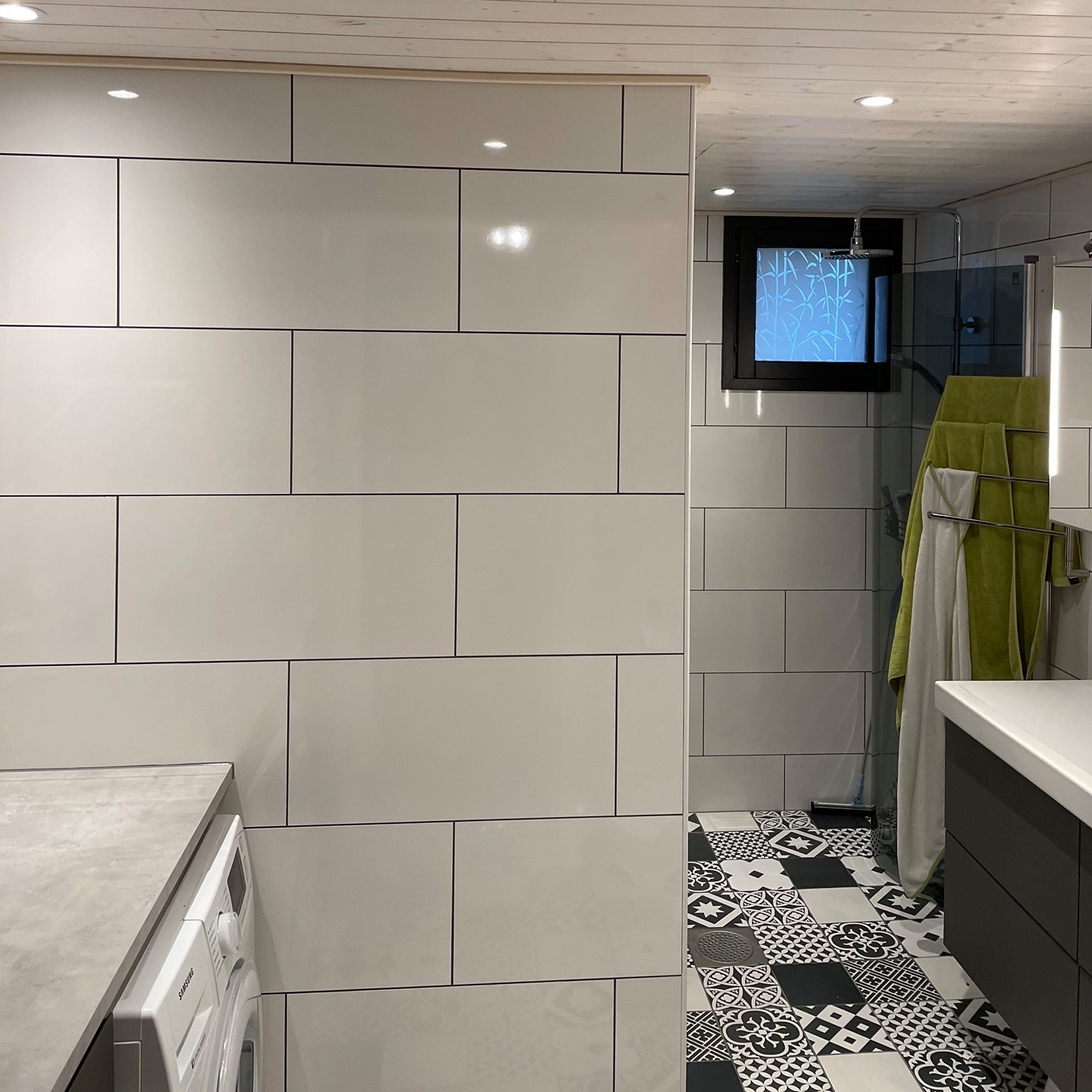 Kylpyhuoneen sisustus, jossa on valkoiset seinälaatat, mustavalkoinen mosaiikkilattia ja puinen kattopaneeli.