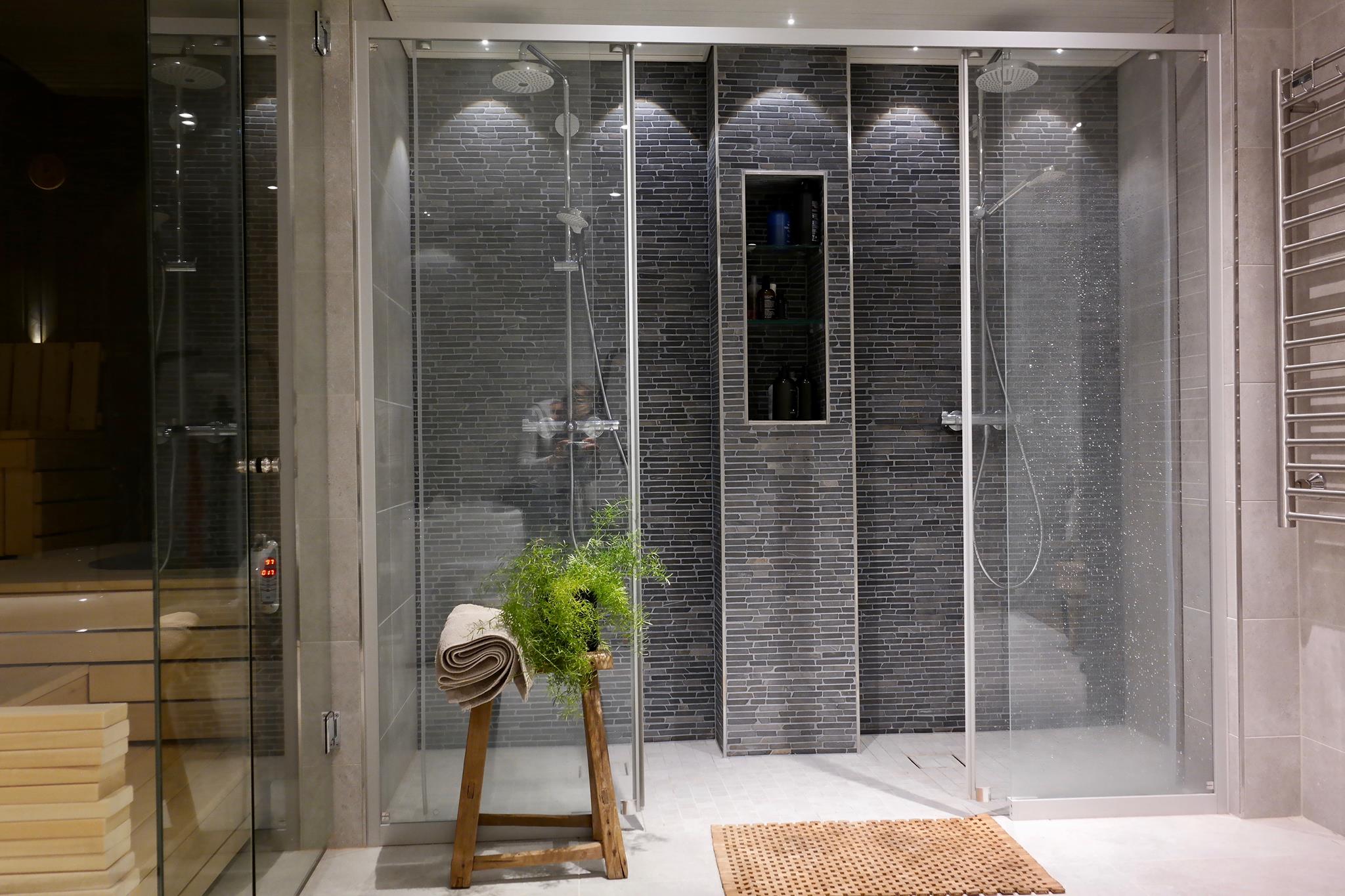 Moderni kylpyhuone, jossa on lasinen suihkukaappi, puinen jakkara pyyhkeellä ja viherkasvilla, sekä tummanharmaata tiiliseinää ja upotettu hylly.
