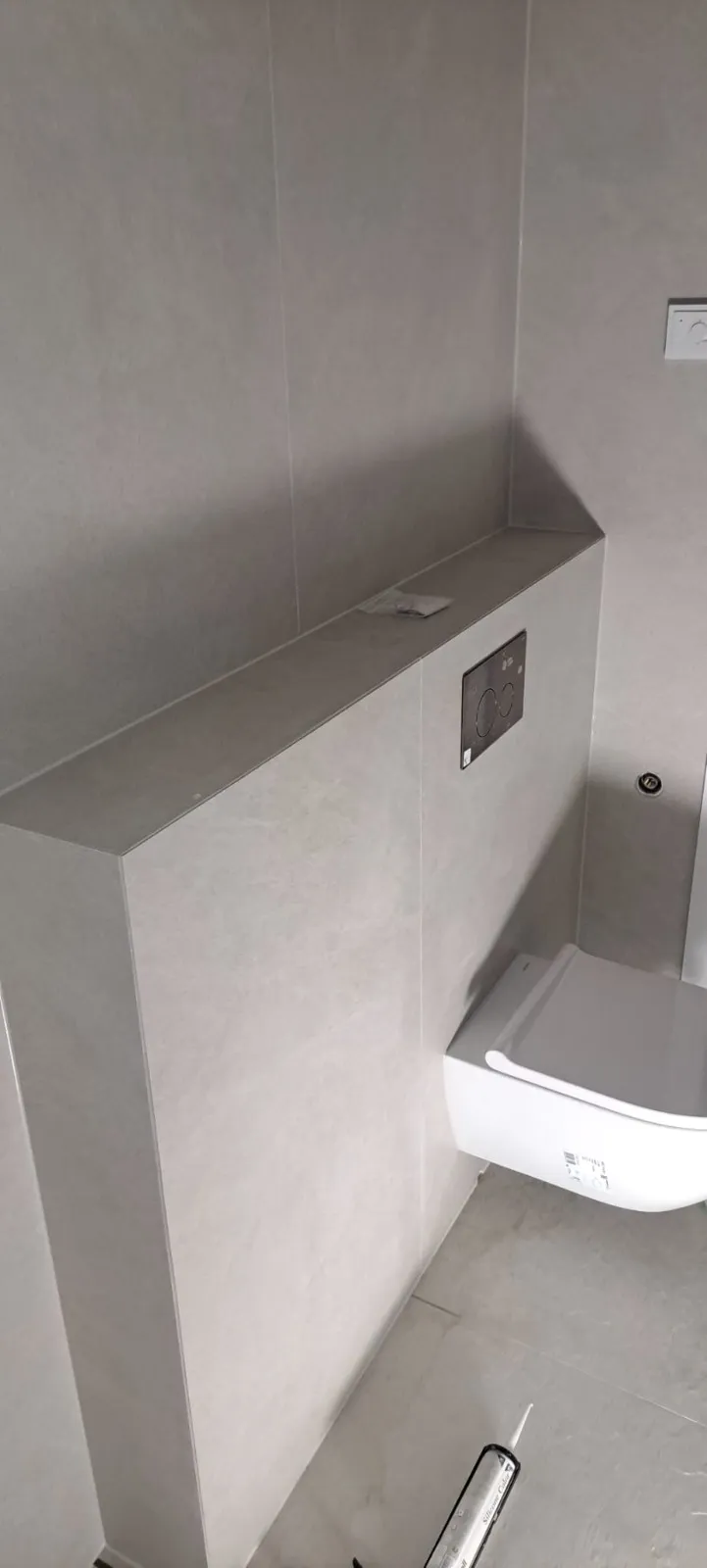 Vaaleanharmaassa kylpyhuoneessa on seinään asennettu WC-istuin, jonka yläpuolella on laatikkomainen rakenne, jossa on painike. Lattialla on silikonipistooli.