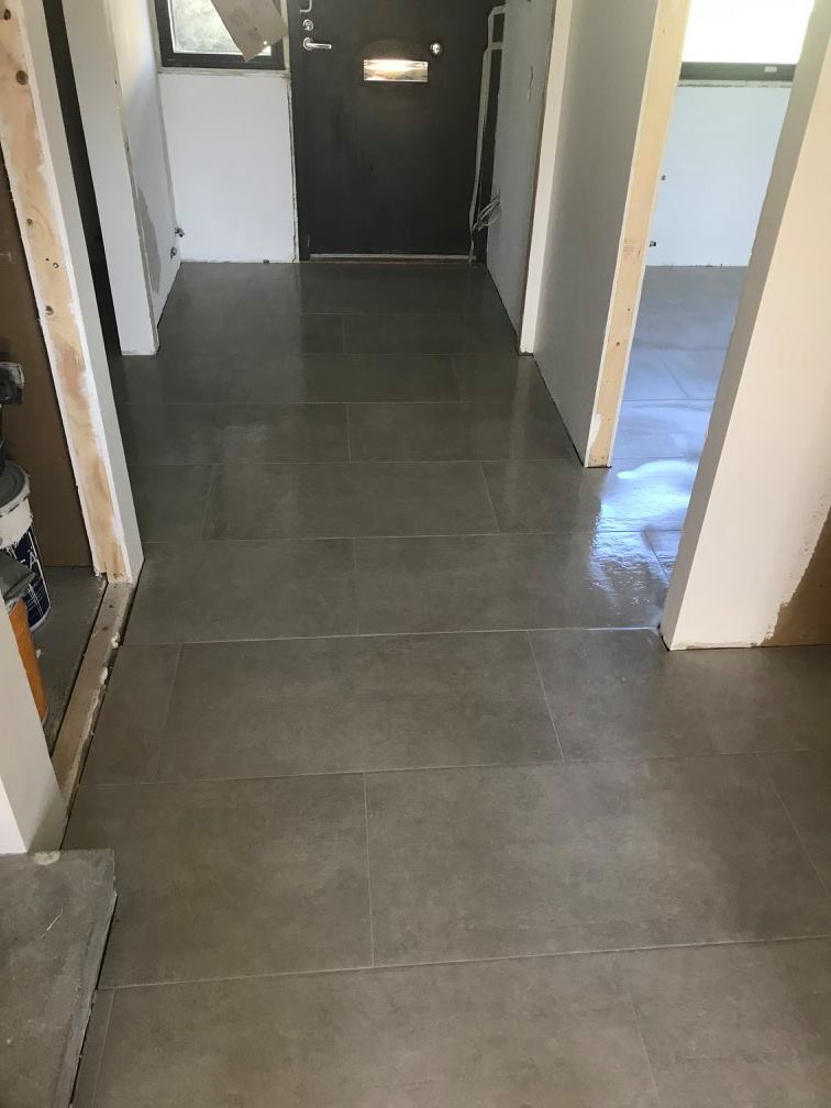 Harmaalla laatoitettu eteinen, jossa näkyy ovi, seinät ja valoisa huone.