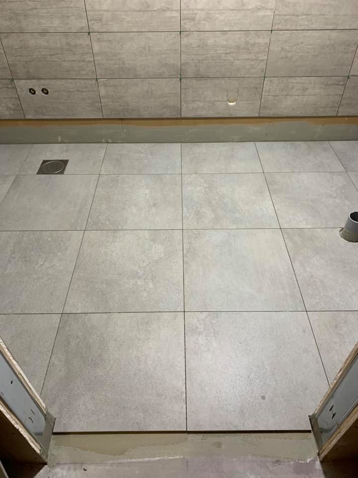 Kuvassa on harmaasävyinen kylpyhuone, jossa on laatoitettu lattia ja seinät. Lattiassa on viemäri ja seinässä on kaksi pientä reikää ja yksi suurempi reikä.