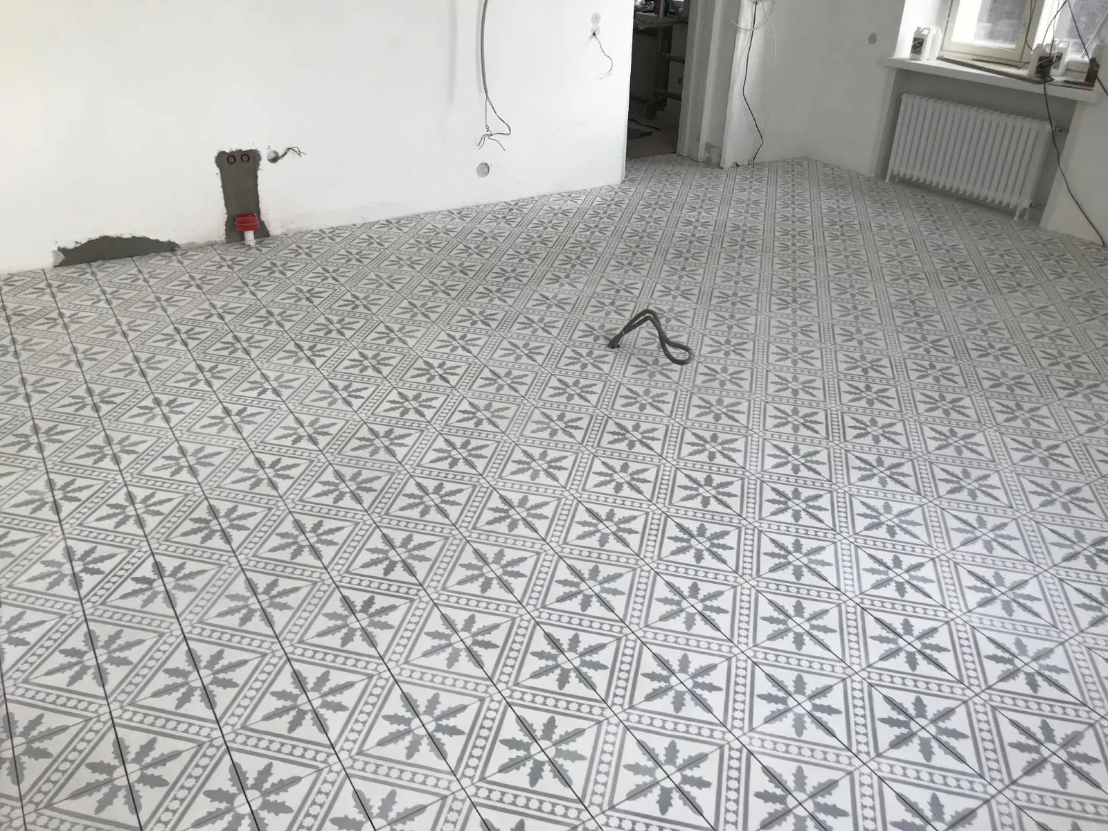 Huoneen lattia on peitetty harmaavalkoisilla, kuvioiduilla laatoilla. Seinät ovat valkoiset ja huoneessa on patteri.