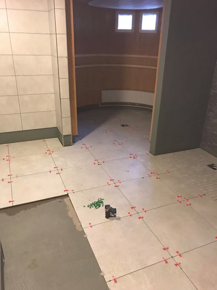 Kuva näyttää laatoitustyön alla olevan kylpyhuoneen, jossa on beigejä lattialaattoja ja punaisia laattaristejä. Taustalla näkyy pyöreä sauna.