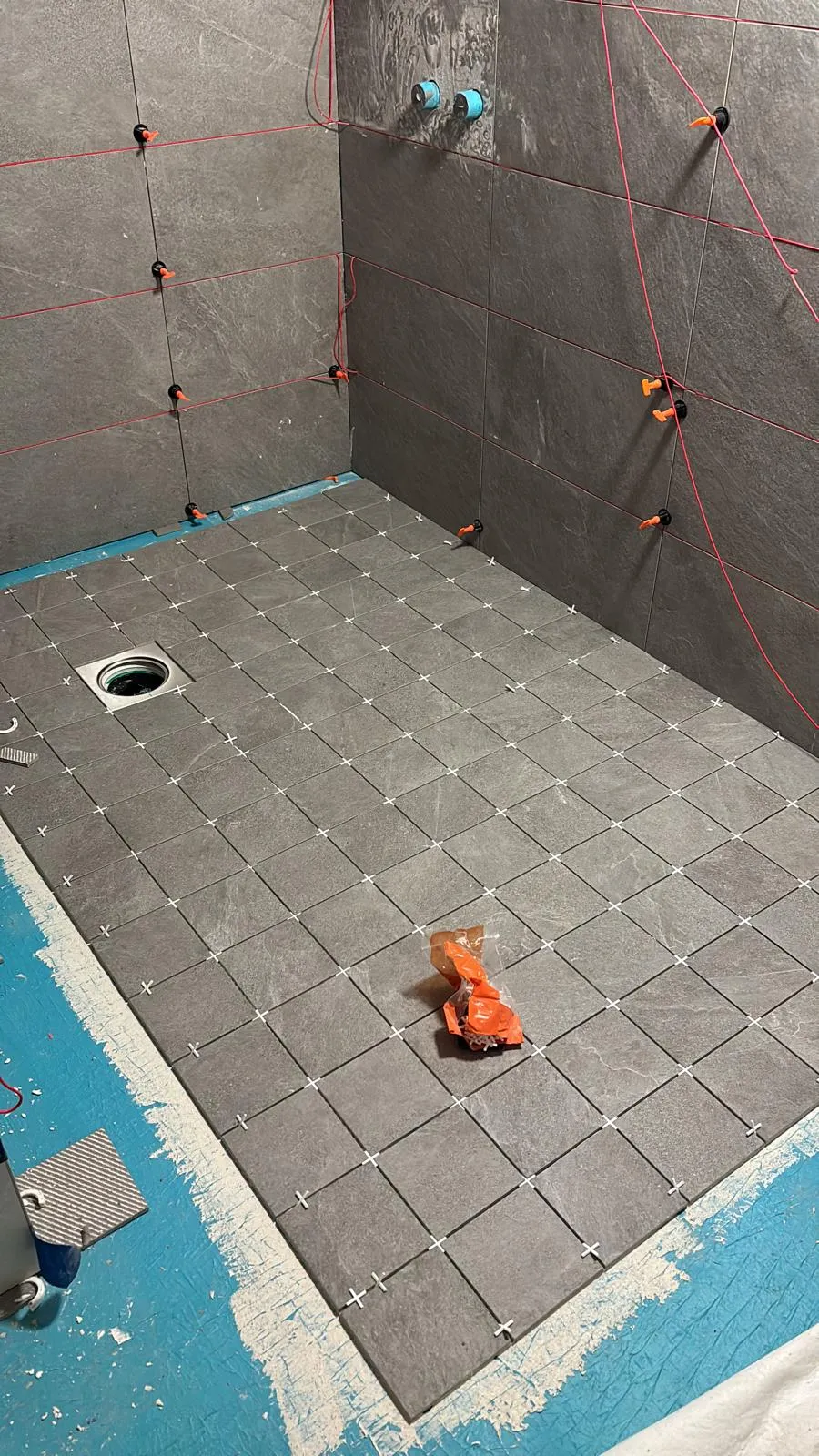 Harmaiden laattojen asennus kylpyhuoneessa, jossa on valkoiset saumat ja oranssi pakkaus lattialla. Punaiset linjat seinillä.