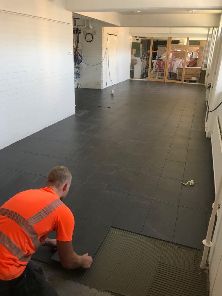 Työntekijä asentaa tummia lattialaattoja valoisassa huoneessa, jossa on valkoiset seinät ja avoin sisäänkäynti.