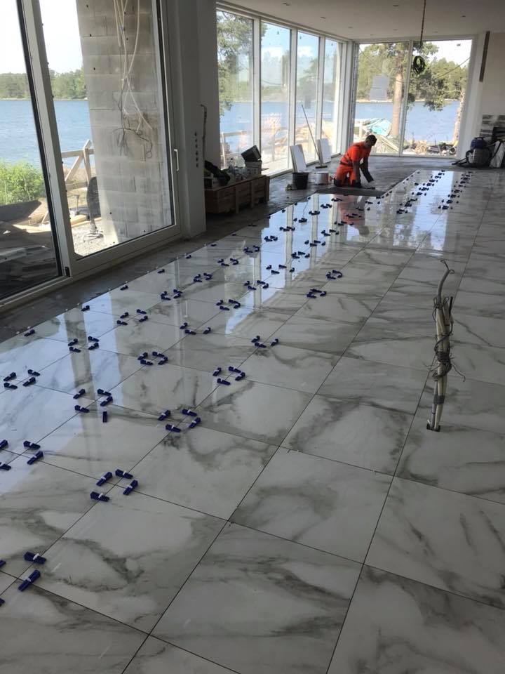 Sisätiloissa on marmorikuvioiset lattialaatat, joissa on sinisiä laattakiiloja, ja työntekijä asentaa laattoja suurten ikkunoiden edessä.