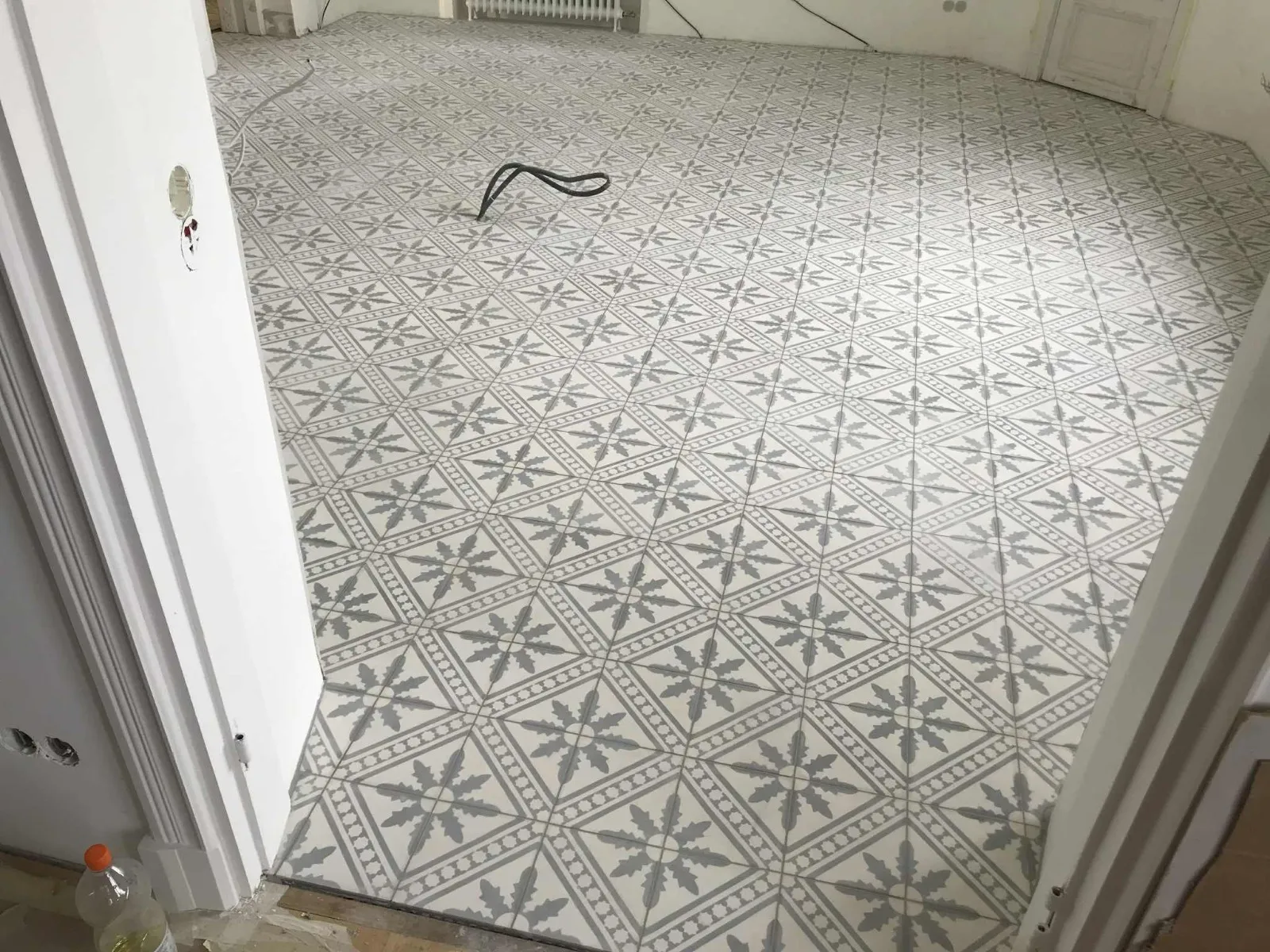 Huoneen lattia on peitetty harmaavalkoisilla, koristeellisilla laatoilla, ja musta johto kulkee lattian poikki.