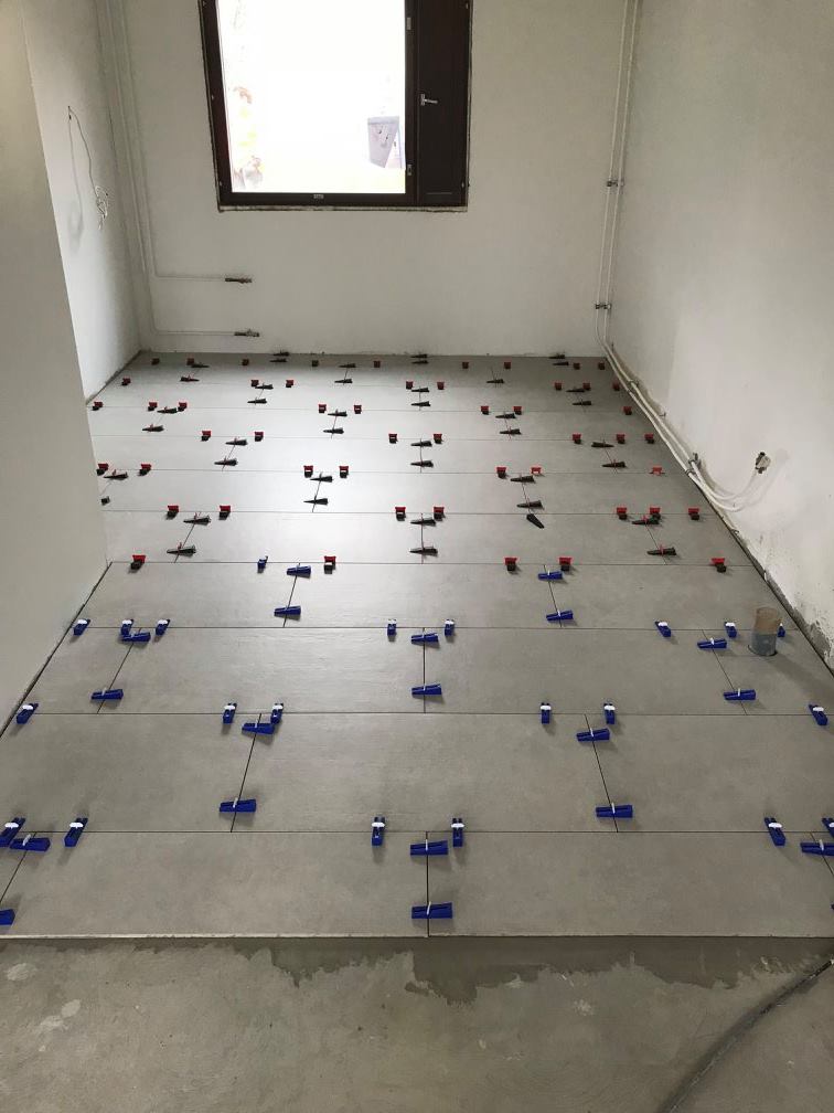 Huoneessa on harmaita lattialaattoja, joissa on sinisiä ja punaisia kiiloja, sekä ikkuna ja valkoiset seinät.