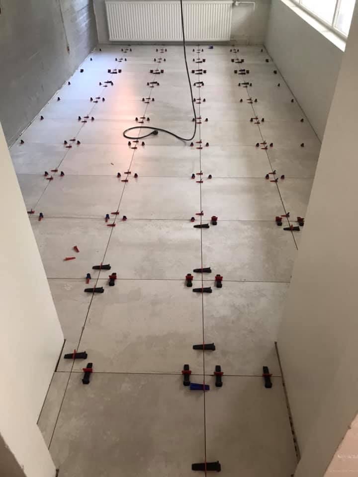 Huoneessa on juuri asennettu vaaleanharmaat lattialaatat, joita pitävät paikoillaan punaiset ja mustat kiilat. Huoneessa on patteri ja ikkuna, ja valaistus on kirkas.