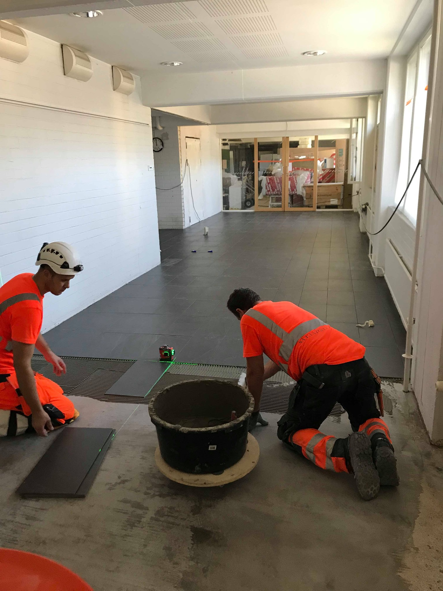 Kaksi rakennusmiestä asentaa lattialaattoja sisätiloissa, käyttäen laseria apuna suorien linjojen saavuttamiseksi, kirkkaassa valaistuksessa.