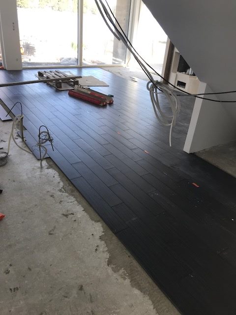 Rakennustyömaa, jossa on tumma puulattia osittain asennettuna, työkaluja ja johtoja lattialla, sekä suuri ikkuna taustalla.