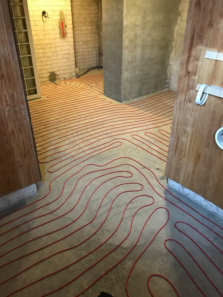 Huoneessa on lattialämmitys punaisilla putkilla, seinät ovat osittain tiiltä ja osittain betonia, valaistus on kirkas.