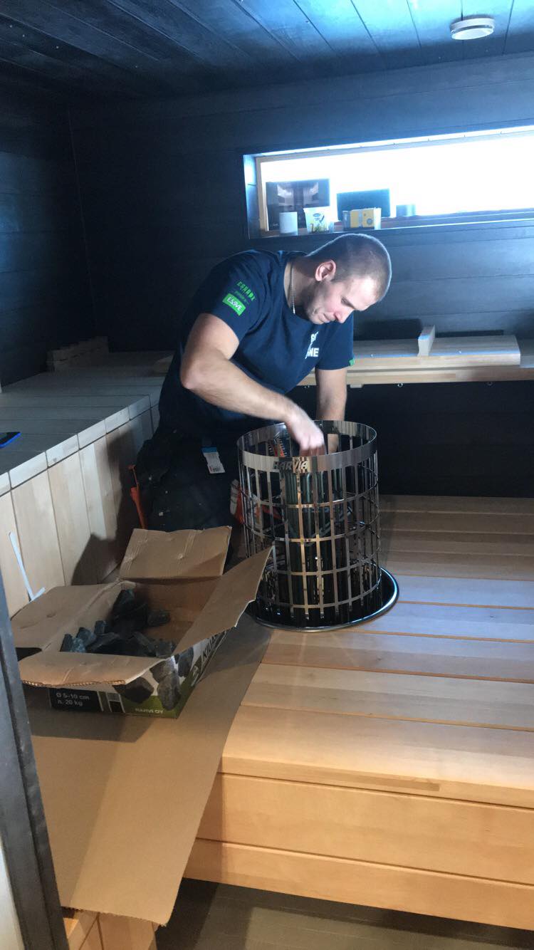 Mies asentaa kiuaskiviä metalliseen kiukaaseen saunassa, jossa on puupanelointi ja ikkuna.