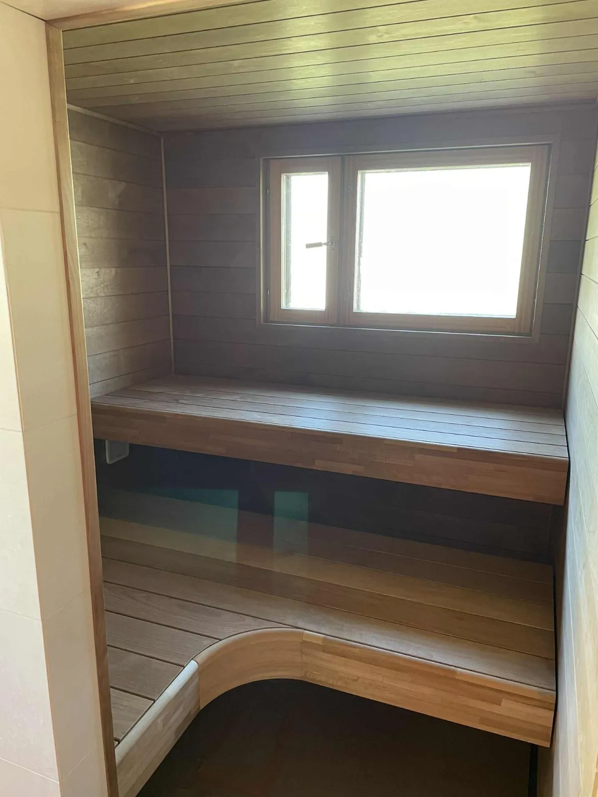 Sauna, jossa on puupanelointi, ikkuna ja puiset penkit. Valoisa ja lämmin tunnelma.
