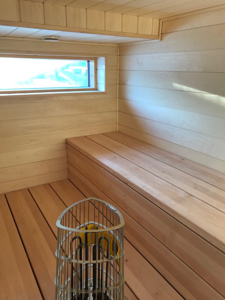 Sauna, jossa on puupanelointi, ikkuna ja kiuas metallisella suojuksella. Vaaleat puunsävyt ja luonnonvalo luovat rauhallisen tunnelman.
