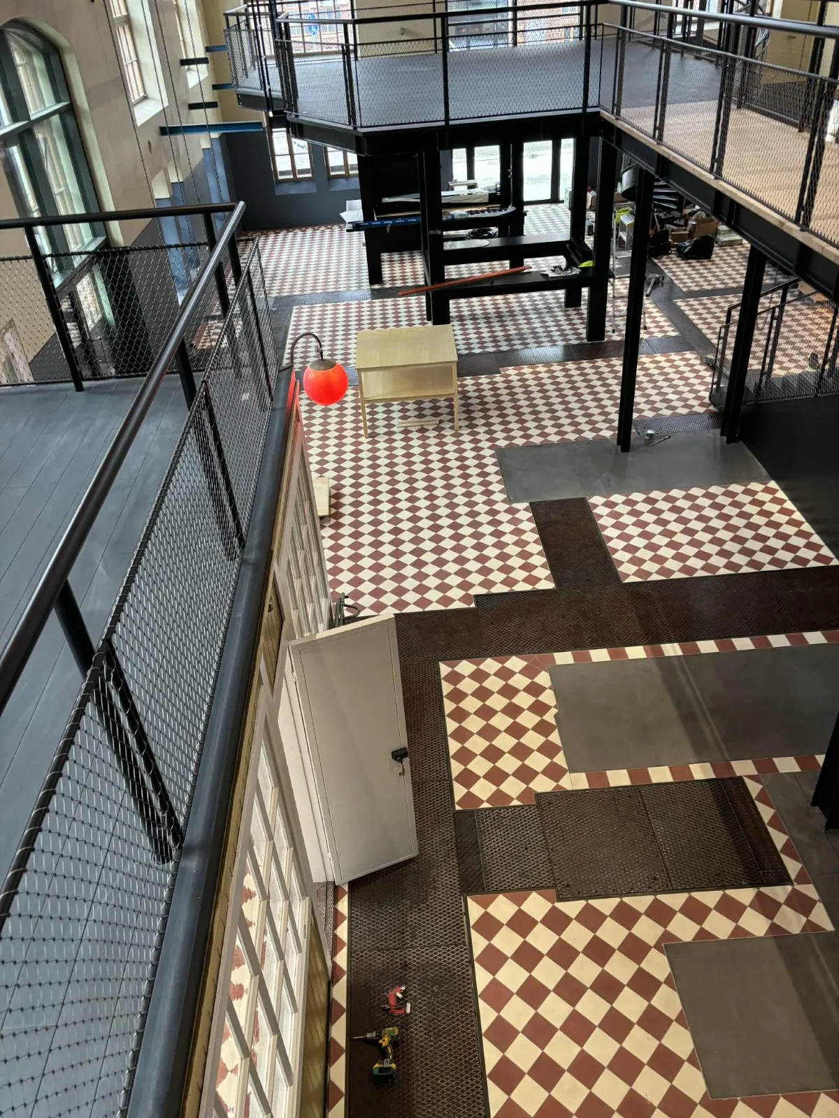 Korkeasta kuvakulmasta otettu kuva sisätilasta, jossa on ruutukuvioinen lattia, metalliset parvekkeet ja valkoinen ovi. Punainen valaisin näkyy myös.