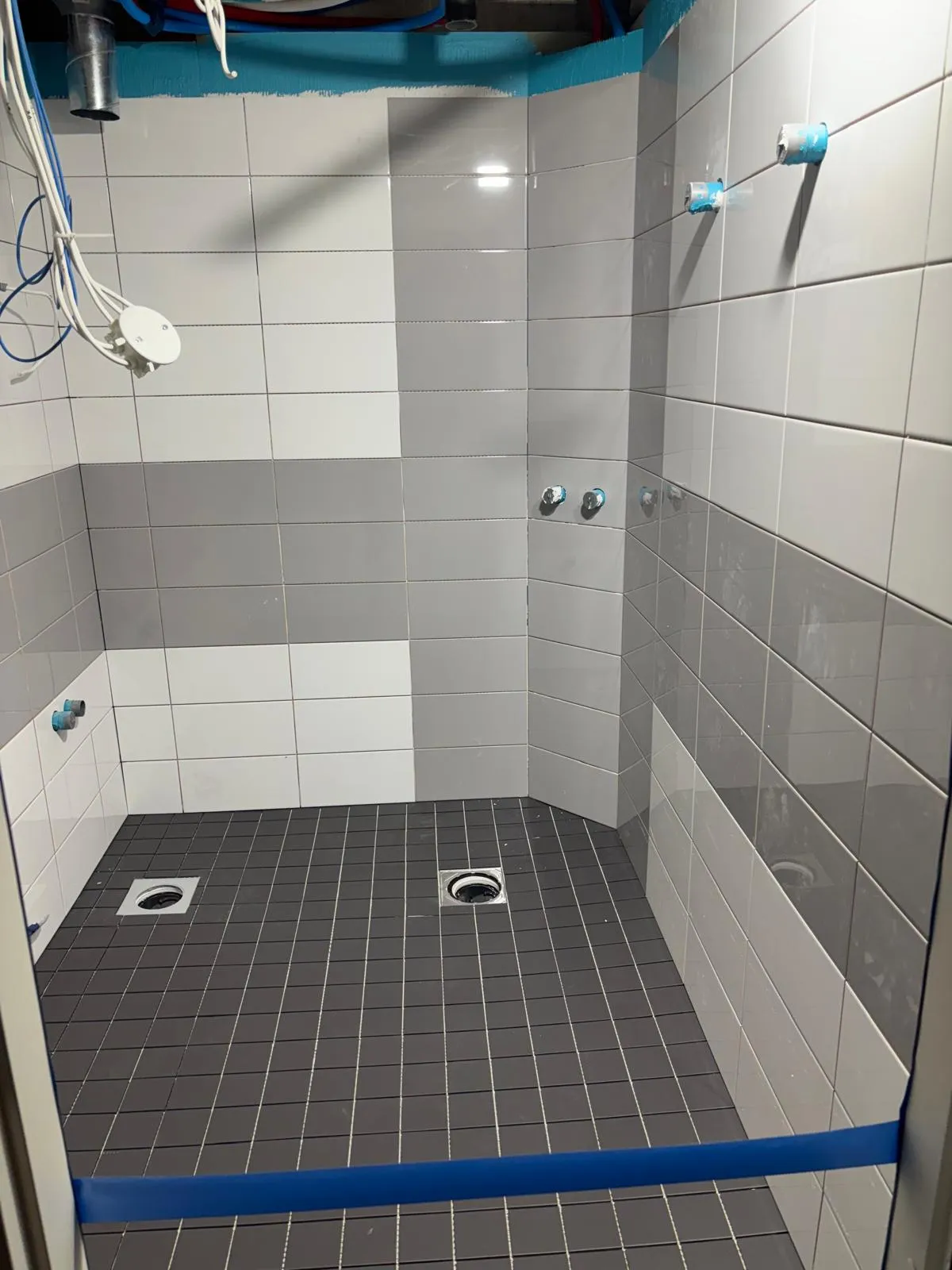 Harmaa ja valkoinen suihkutila, jossa on tummat lattialaatat ja siniset putket katossa.