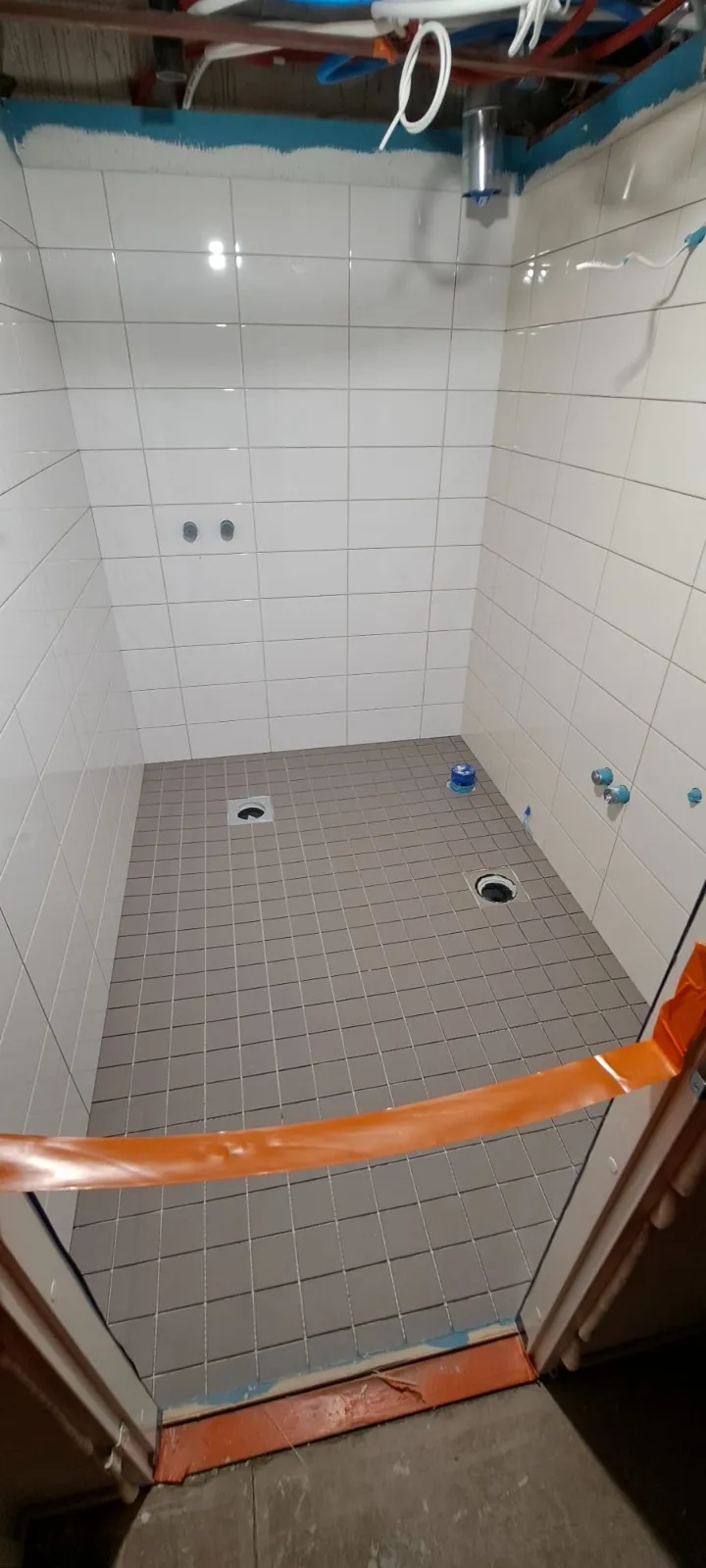 Valkoisilla seinälaatoilla ja harmailla lattialaatoilla varustettu suihkutila, jossa on kaksi lattiakaivoa ja oranssi suojateippi.