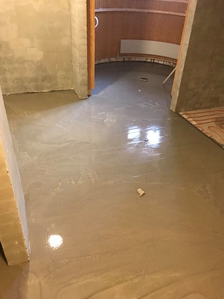 Valokuva huoneesta, jossa on juuri tasoitettu lattia, joka heijastaa valoa. Seinät ovat osittain rapattuja ja osittain puupaneloituja.