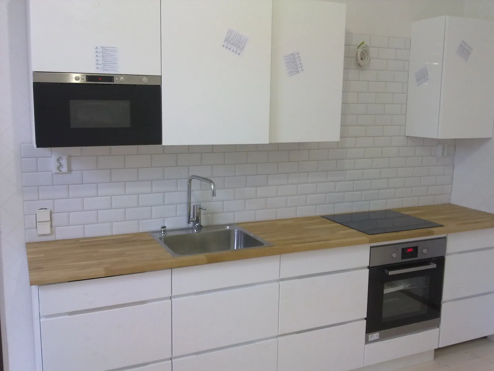 Valkoinen keittiö, jossa on puinen työtaso, valkoiset kaapit ja laattaseinä.